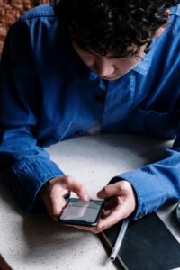 A young man in a blue shirt uses his smartphone in a cozy café, focusing on typing messages.