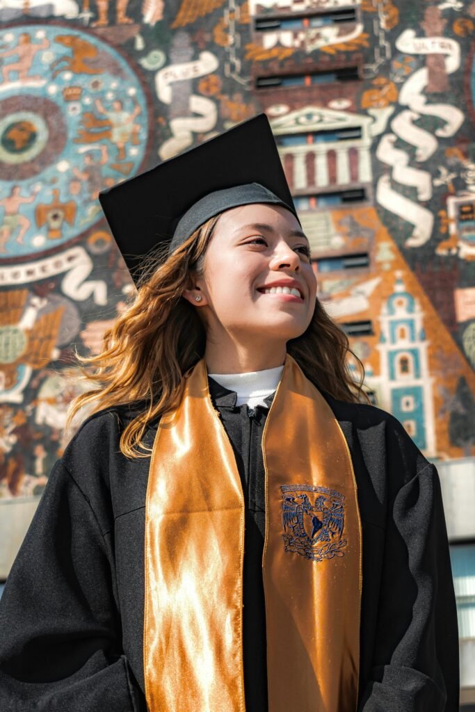A proud young woman in a graduation gown smiles against a vibrant mural backdrop.