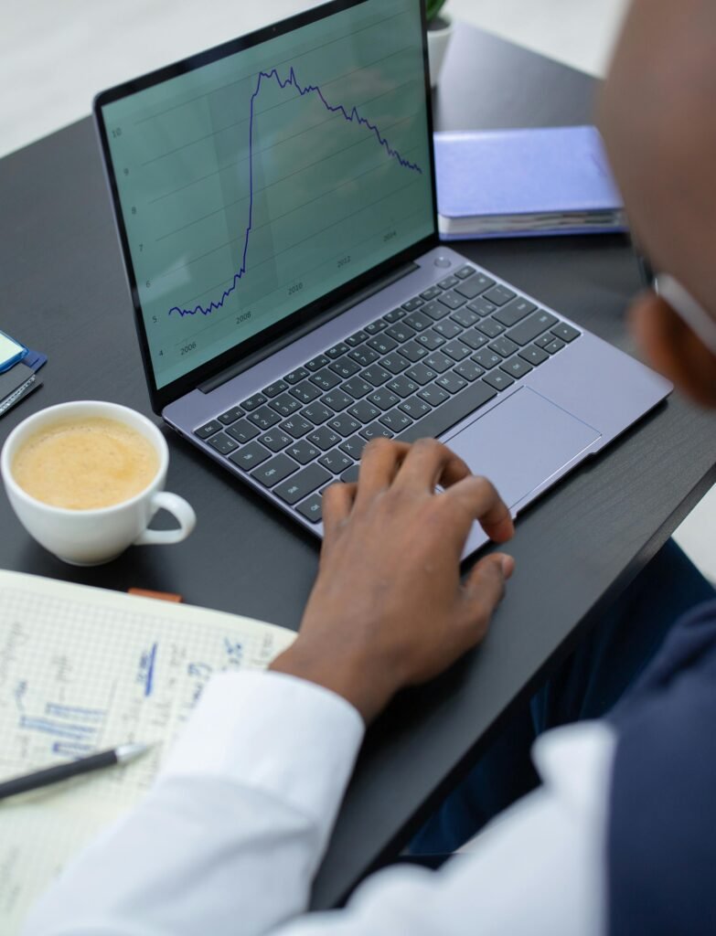 Professional analyzing financial charts on a laptop with notebook and coffee in office setting.