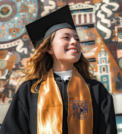 A proud young woman in a graduation gown smiles against a vibrant mural backdrop.