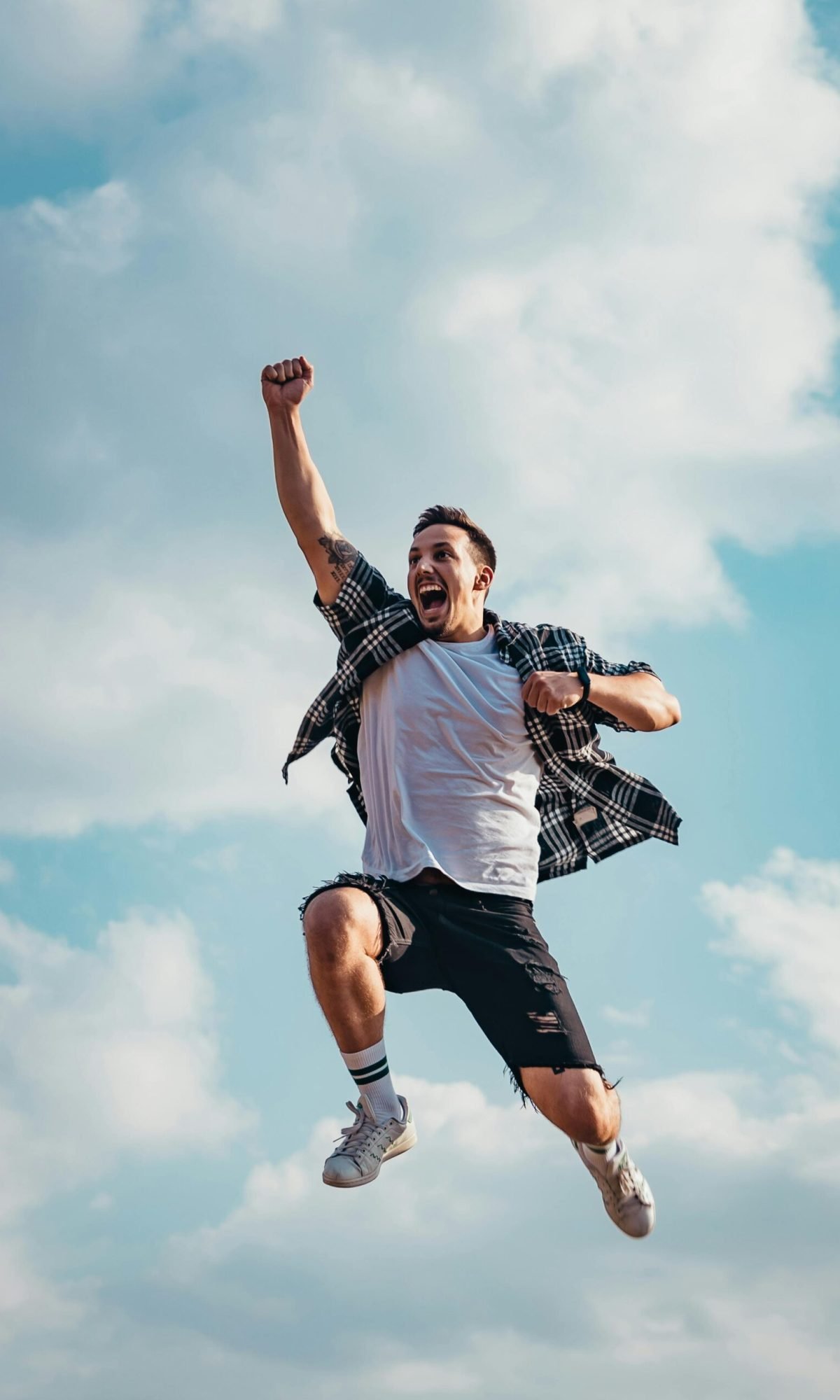 A joyful young man jumps midair with clouds and blue sky in the background, exuding energy and freedom.