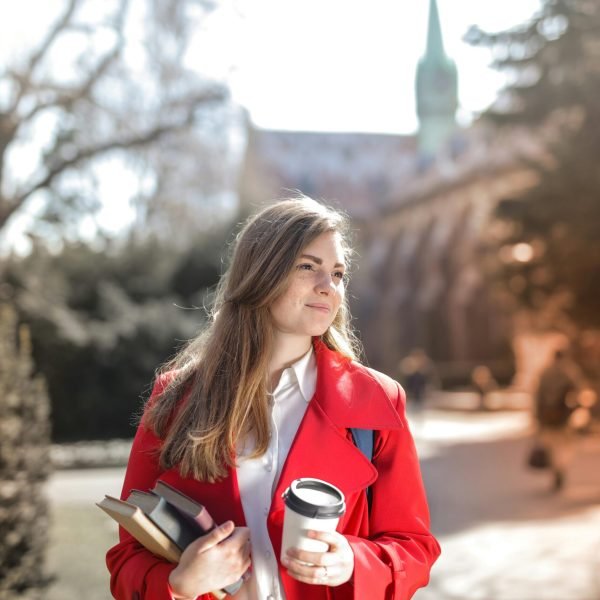 pexels-photo-3755760-3755760 Smiling woman in red coat with coffee and books, enjoying leisure time outdoors on a university campus.