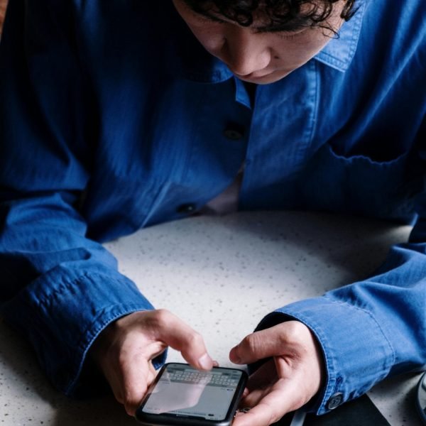 A young man in a blue shirt uses his smartphone in a cozy café, focusing on typing messages.