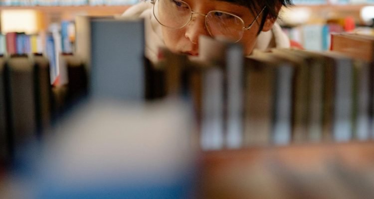 Asian man with eyeglasses looking at books on a library shelf