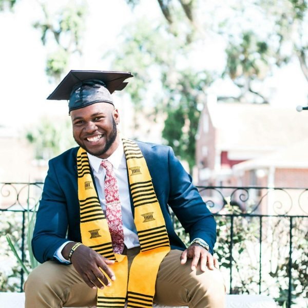 Smiling graduate sitting outside wearing a cap and gown with stole.